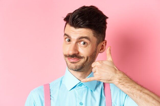 Close-up of handsome young guy with moustache, showing phone gesture, asking to call him, standing on pink background.