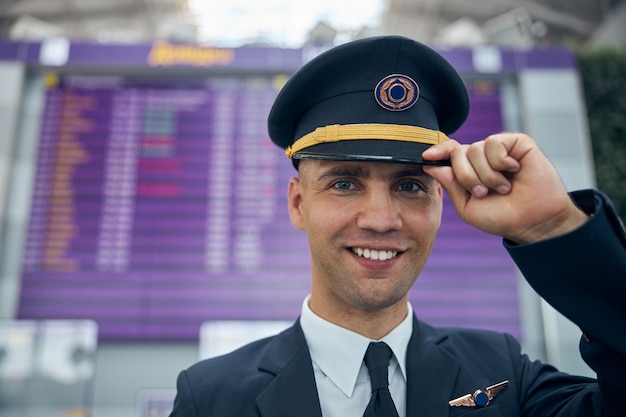 Close up of handsome young man airline captain touching his pilot hat and smiling