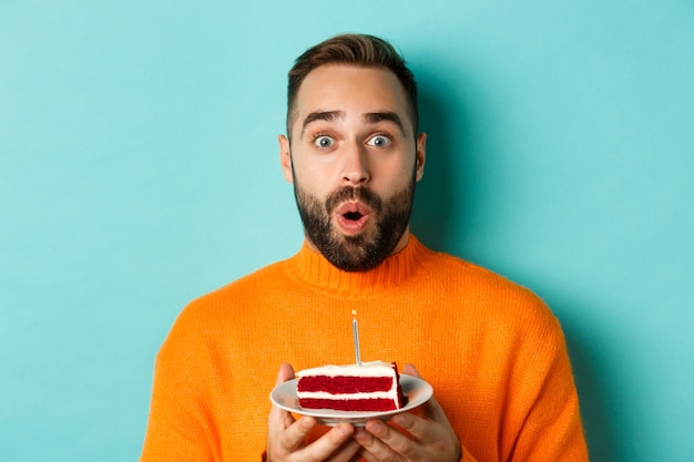 Close-up of happy adult man celebrating birthday, holding bday cake with candle and making wish, standing against turquoise background.