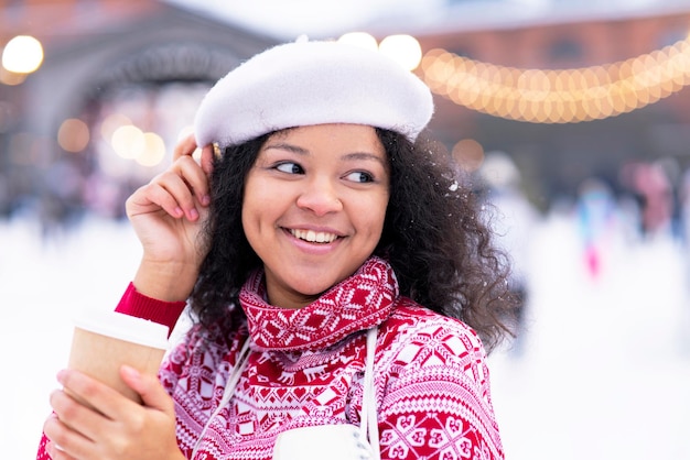 Close-up of a happy African American girl drinking coffee from a glass on a walk in winter. Portrait of a young woman. Dark-skinned black woman in the park.