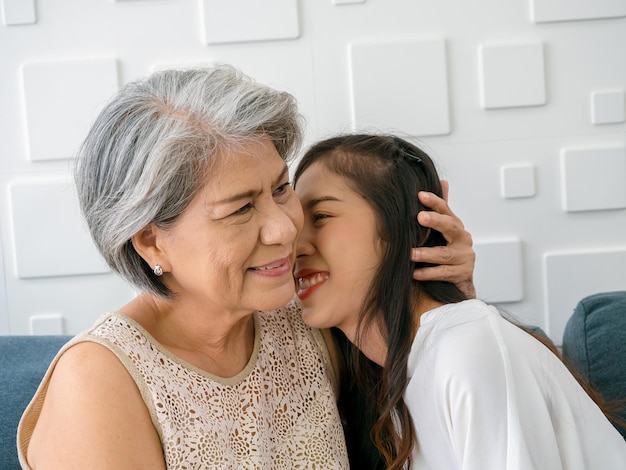 Close up happy Asian senior mother white hair embracing her beautiful daughter while she kiss her cheek with love care and smile while sit on sofa on white background in living room at home