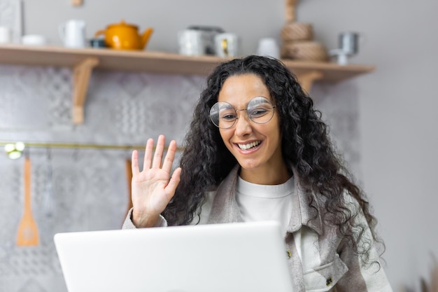 Close up of happy hispanic woman talking on video call at home smiling and waving greeting gesture