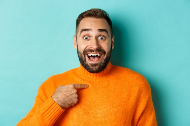 Close-up of happy young man pointing at himself with surprise and excitement, standing over light blue background.