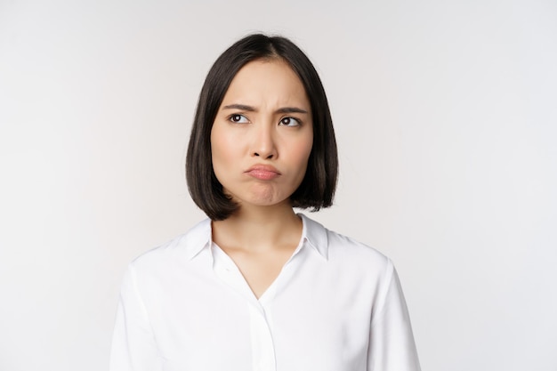 Close up head portrait of young asian woman looking upset and disappointed at left copy space grimacing and frowning displeased white background