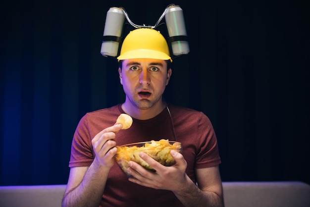 Close up of the hipster man with beer helmet on the head eating chips while watching tv at home