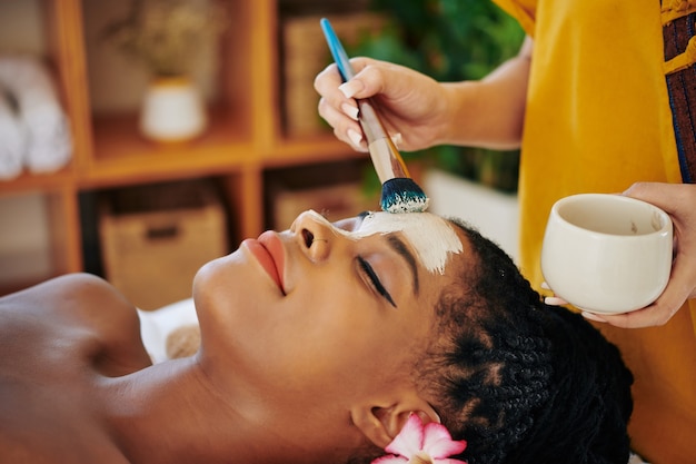 Close-up image of beautician applying purifying and minimizing pores clay mask on face of beautiful young Black woman