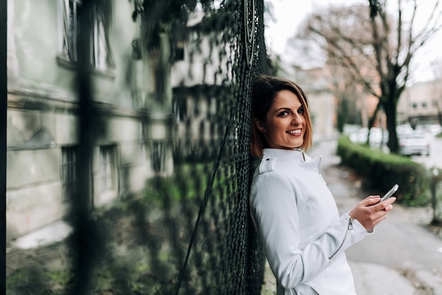 Close-up image of a beautiful girl using phone while standing at city street, leaning on the fence.