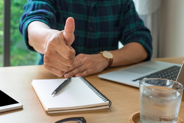 Close up image of businessman showing thumbs up on office desk with laptop smart phone eyeglasses notebook pen He was delighted and felt that today's work was a great day