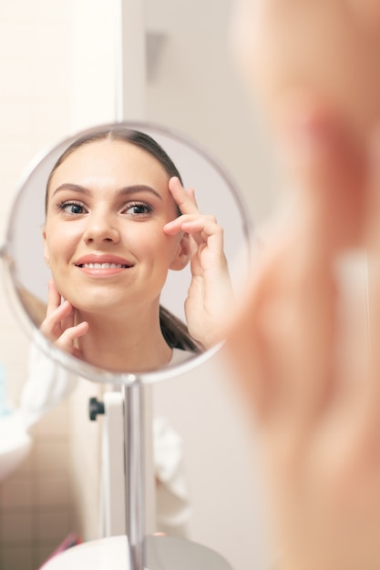 Close up image of the cheerful emotional young lady smiling and touching the skin on her face while looking at the reflection in the mirror