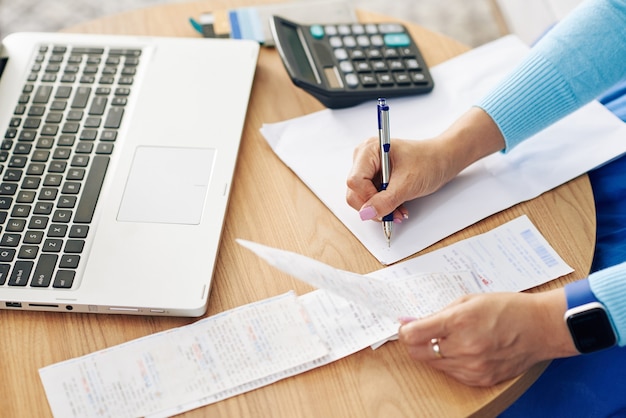 Close-up image of female entrepreneur checking bills and writing figures on blank sheet