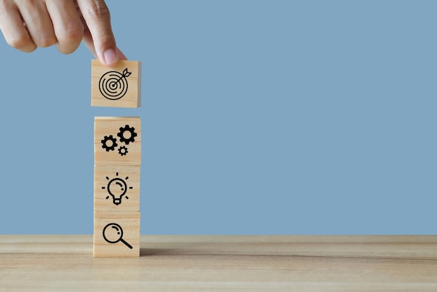 Close up image of hand holding wood cube block with business icon target idea magnifying glass gear and placed it on office desk