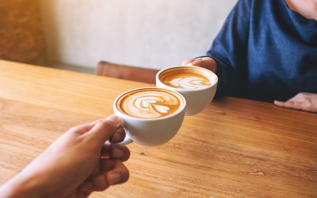 Close up image of a man and a woman clinking two coffee mugs on wooden table in cafe