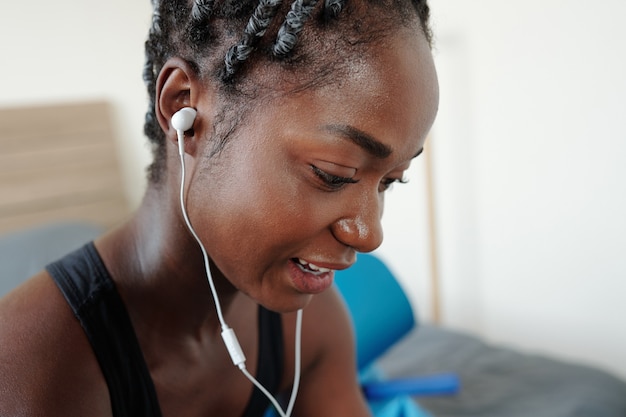 Close-up image of young Black woman sweaty after training at home choosing next song on smartphone or player