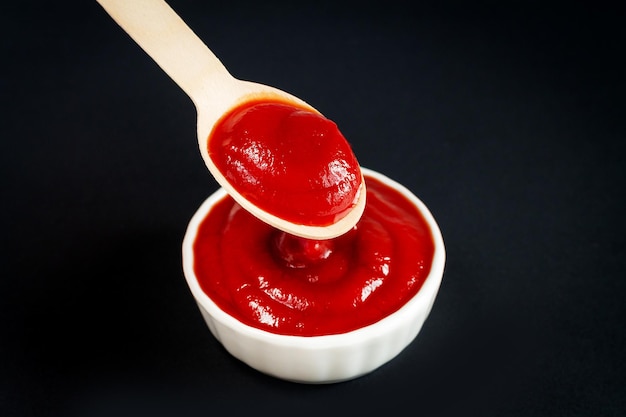 Close-up of ketchup or red tomato sauce in wooden spoon and white bowl on black background