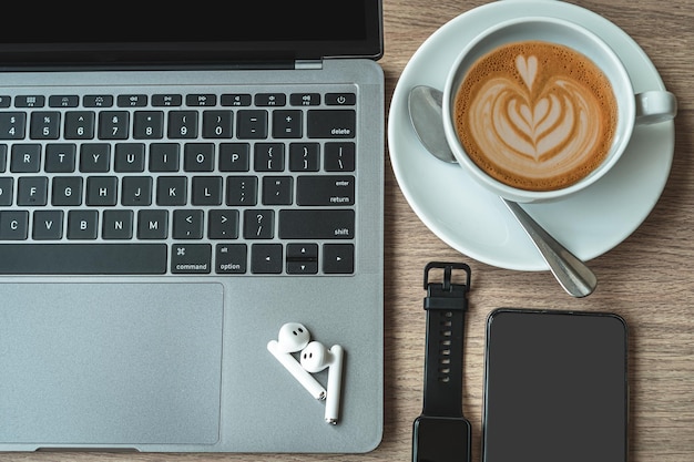 Photo close-up of keyboard laptop computer, wireless earphones focus with smartwatch,hot coffee in cup mug and smartphone empty screen on wooden background office desk in coffee shop like the background