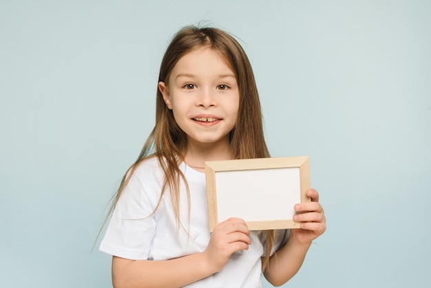 Close up little girl holding white blank frame posing isolated on pastel blue background in studio Copy space mockup