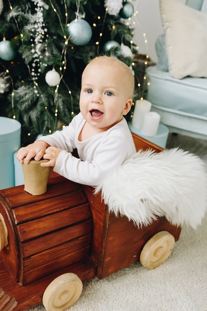 Close up of a lovely Caucasian baby sitting in a wooden toy train near decorated Christmas Tree