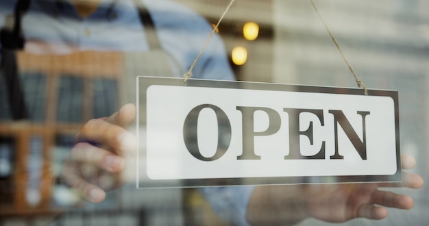 Close up of the male Caucasian hands turning a signboard on the glass door of the shop from CLOSED to OPEN and man in glasses looking out on the street.