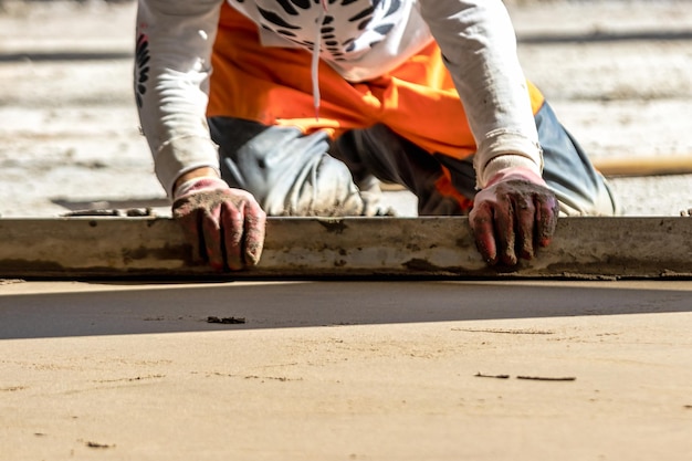 Close up of man builder placing screed rail on the floor covered with sandcement mix at construction site Male worker leveling surface with straight edge while screeding floor Blurred background