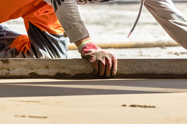 Close up of man builder placing screed rail on the floor covered with sandcement mix at construction site Male worker leveling surface with straight edge while screeding floor Blurred background