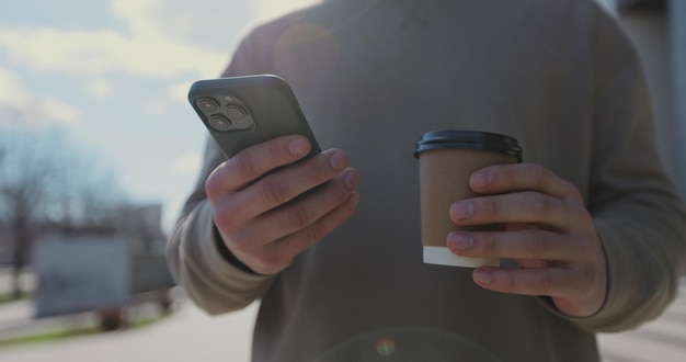 Close up of a man drinking to go coffee and browsing smartphone social networking at urban modern street People and communication concept