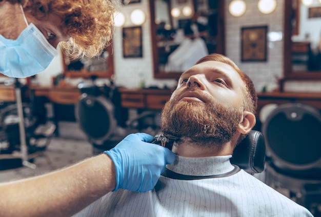 Close up man getting hair cut at the barbershop wearing mask during coronavirus pandemic. Professional barber wearing gloves. Covid-19, beauty, selfcare, style, healthcare and medicine concept.