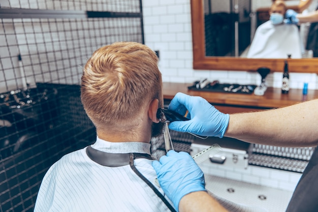 Close up man getting hair cut at the barbershop wearing mask during coronavirus pandemic. Professional barber wearing gloves. Covid-19, beauty, selfcare, style, healthcare and medicine concept.