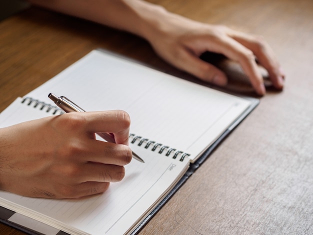 Close up of man's hands writing in notebook placed on wooden desktop