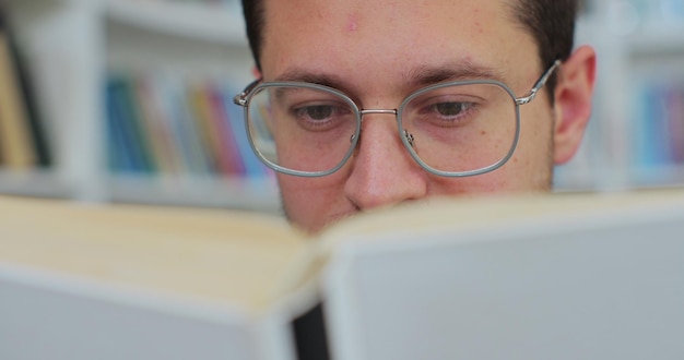 Close up man student in glasses is spending his time in library examining a book and reading the contents Looking at the camera Student lifestyle education concept