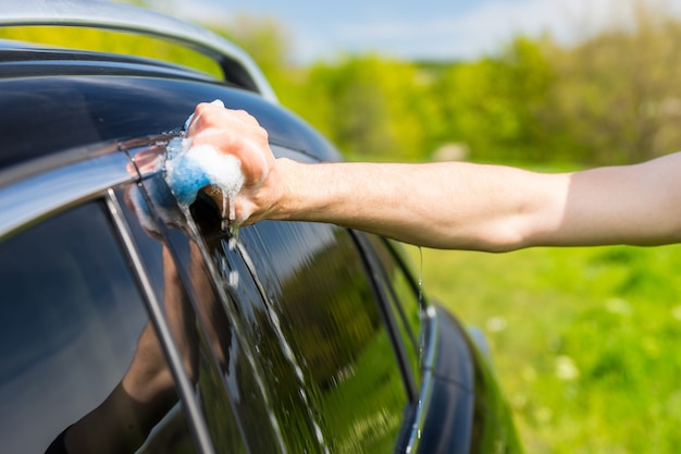 Close Up of Man Washing Windows of Black Luxury Vehicle with Soapy Sponge in Green Field on Bright Sunny Day