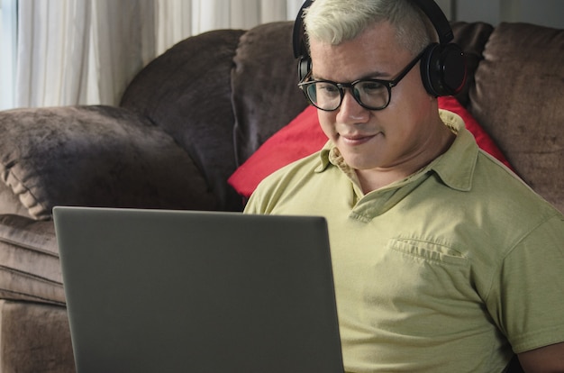 Close Up man with glasses sitting on the living room floor next to the sofa, checking laptop with headphones