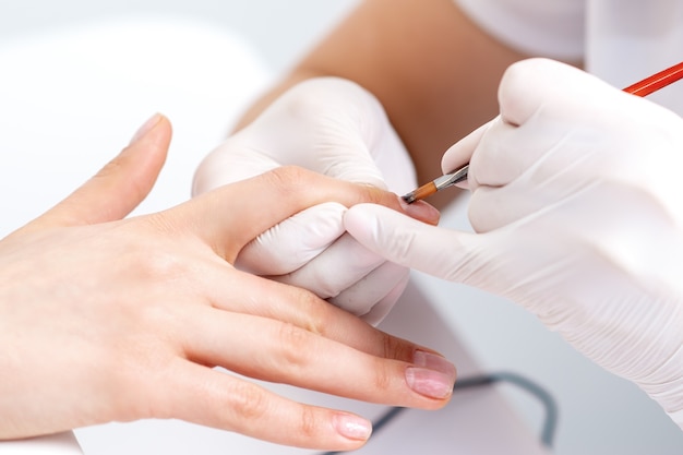 Close up of manicure master in rubber gloves applying transparent nail polish on female nails in beauty salon