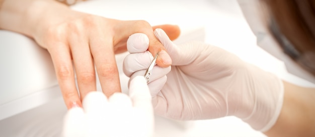 Close up of manicure master with manicure scissors removes cuticles of female nails at beauty salon.