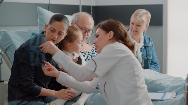 Close up of medic explaining x ray results to woman in hospital ward, talking about diagnosis of senior man. Doctor holding radiography in front of daughter, niece and wife of patient.