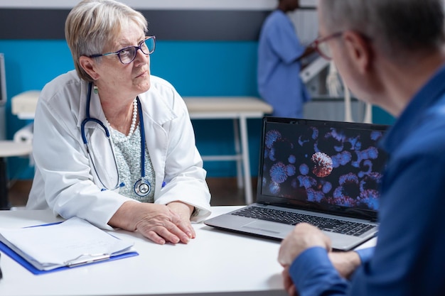 Close up of medic and patient discussing about virus bacteria illustration on laptop. General practitioner using computer to explain visual representation of covid 19 bacteria to senior man.