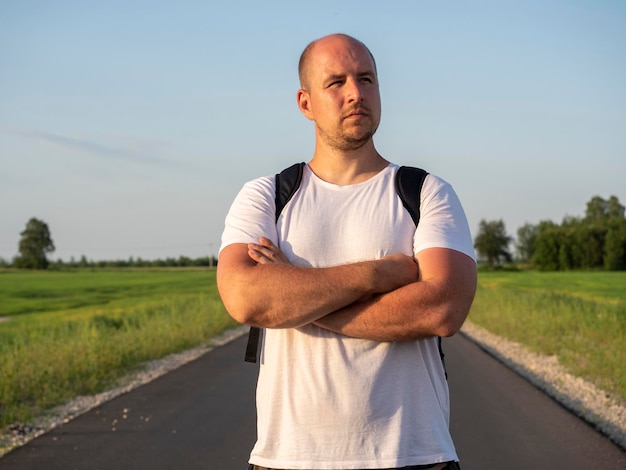 Close up a middleaged man with a backpack on his back is facing the camera on the road in the field His hands are folded on his chest