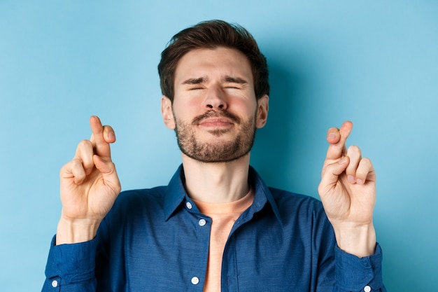 Close-up of modern young man making wish, close eyes and cross fingers for good luck, praying for dream come true, standing on blue background.