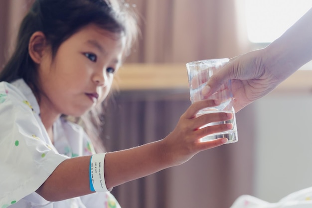 Close up on mother hand giving a glass of water to her sick daughter. Sick asian child drinking water after taking medicine when stay in private patient rooms of hospital.