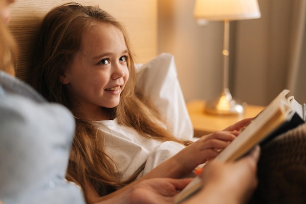 Close-up of mother and smiling adorable daughter reading together children book before going to sleep while lying in bed in nursery bedroom, near lamp. Concept of family leisure activity at home.