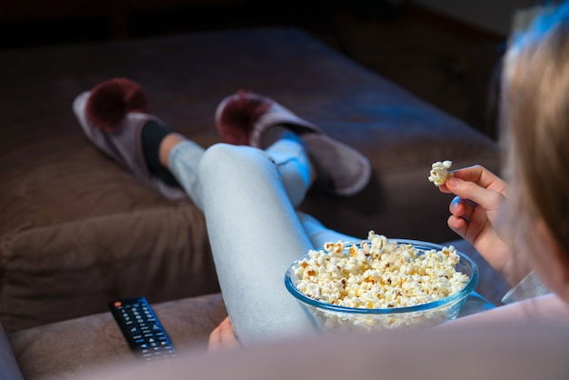 Close up o f hand waking popcorn from a bowl while watching TV. Person sitting in comfortable couch and watching home cinema in the dark.