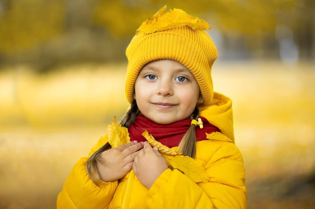 Close up outdoor portrait of cute little girl, wearing yellow jacket,cap, red scarf, posing with smile at camera on the background of autumn trees. Yellow fallen leaves on girl's head and shoulders.