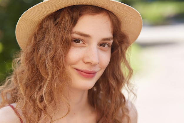 Close up outdoor portrait of young attractive woman wearing straw hat, has foxy wavy hair, looking directly , has charming smile