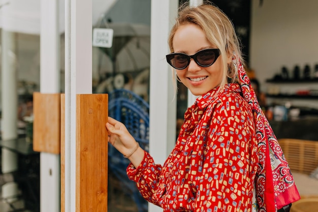 Close up outside portrait of attractive happy girl with blond hair wearing bright summer dress and sunglasses posing at camera while leaving cafeteria in warm summer day