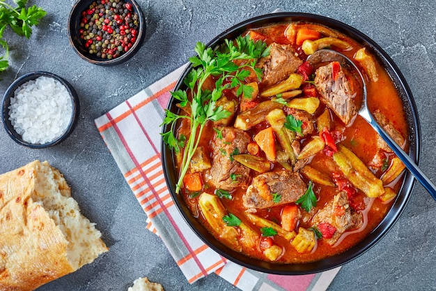 Close-up of overhead view of delicious Beef and Okra Stew served in a bowl with spoon, on an grey concrete table with ciabatta bread, flat lay