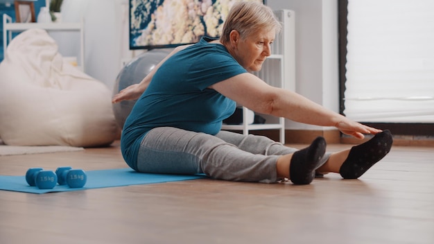 Close up of pensioner doing physical exercise to stretch muscles at home. Retired woman stretching arms and legs while sitting on yoga mat. Senior person training with fitness activity