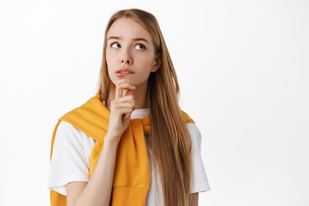 Close up of pensive young woman, thinking and looking at upper right corner copy space, making choice, deciding something, standing against white wall