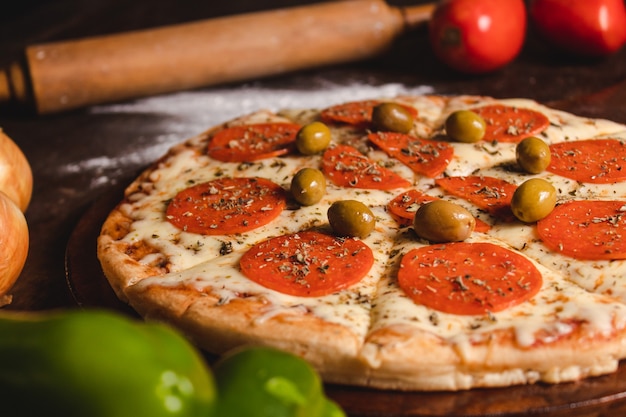 Close-up of a pepperoni pizza with olives and different vegetables around it on top of a wooden table.