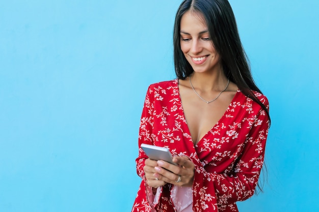 Close up photo of an attractive dark-haired girl in a red dress with floral pattern, posing with a smartphone in her hands, smiling while reading something