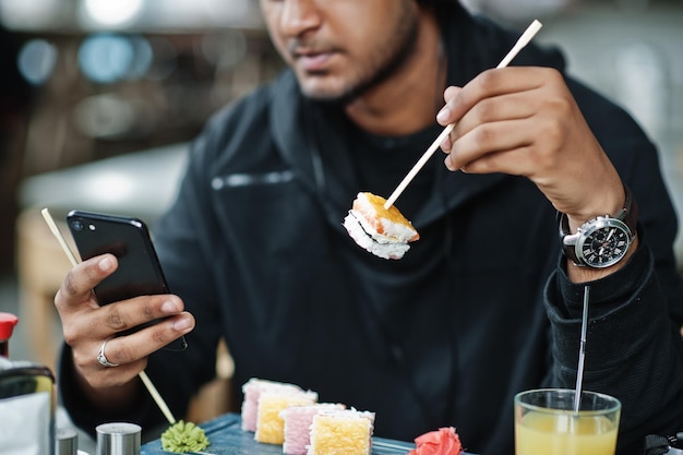 Photo close up photo of casual and stylish young asian man at cafe eating sushi and looking at mobile phone