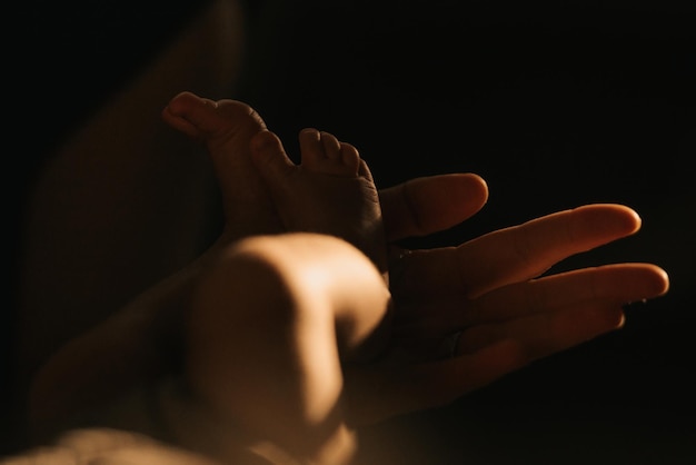 A close-up photo of the gentle palm of the mother who is holding the heels of her newborn baby. The legs of the infant in the sunlight in the evening.